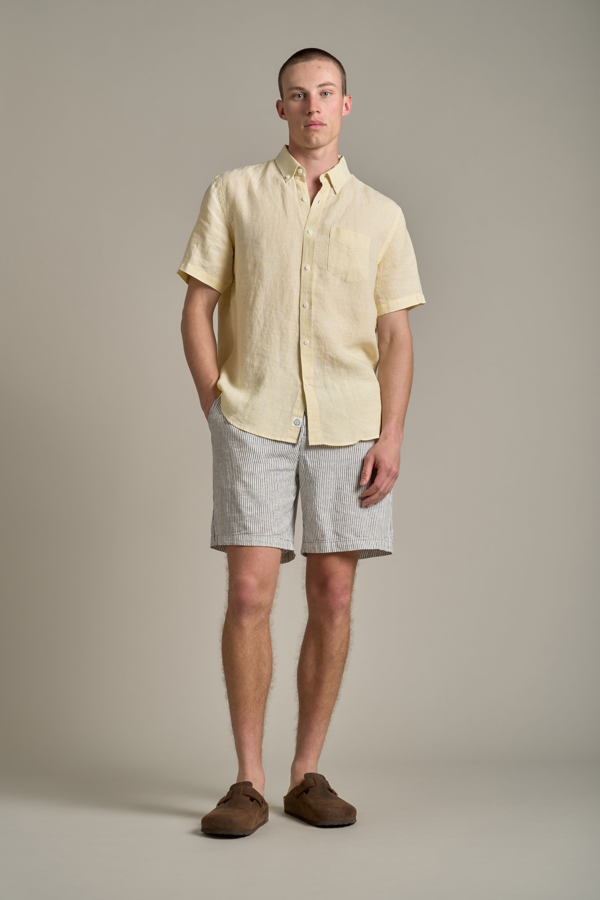 A man stands against a plain background wearing the Barkers Gentry Linen Shirt in light yellow, light gray striped shorts, and brown loafers, hands in pockets—a breathable choice for any summer wardrobe.