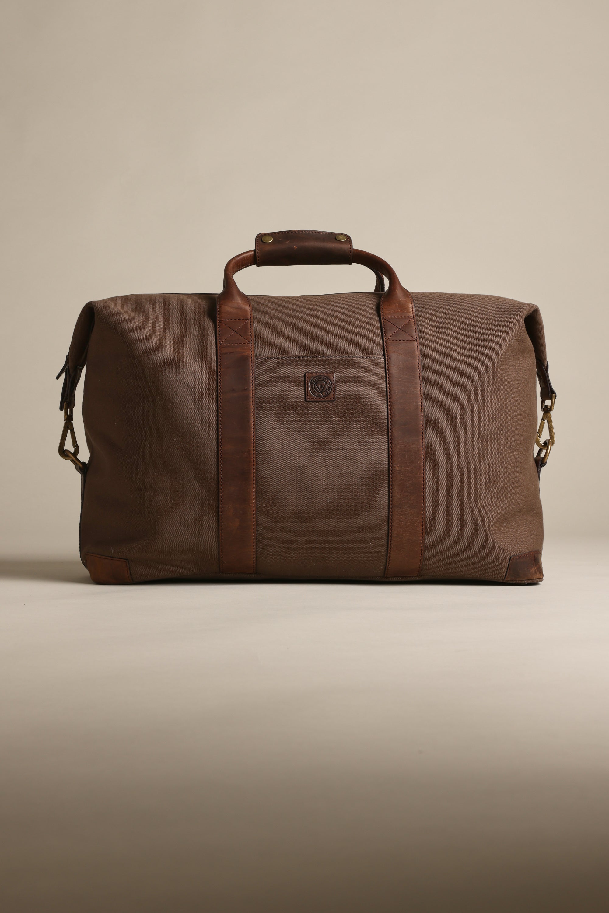 The Barkers Newman Canvas Weekender, a spacious brown men's bag with leather handles, accents, a front square logo, and sturdy metal hardware, stands upright against a neutral background.