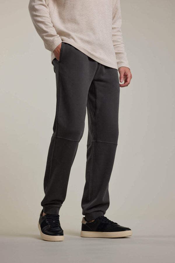 A person wears a beige long-sleeve shirt and Barkers Newton GD Track Pant in a vintage wash, standing with one hand in their pocket. They're also wearing black sneakers with white soles. The photo is cropped at the shoulders.