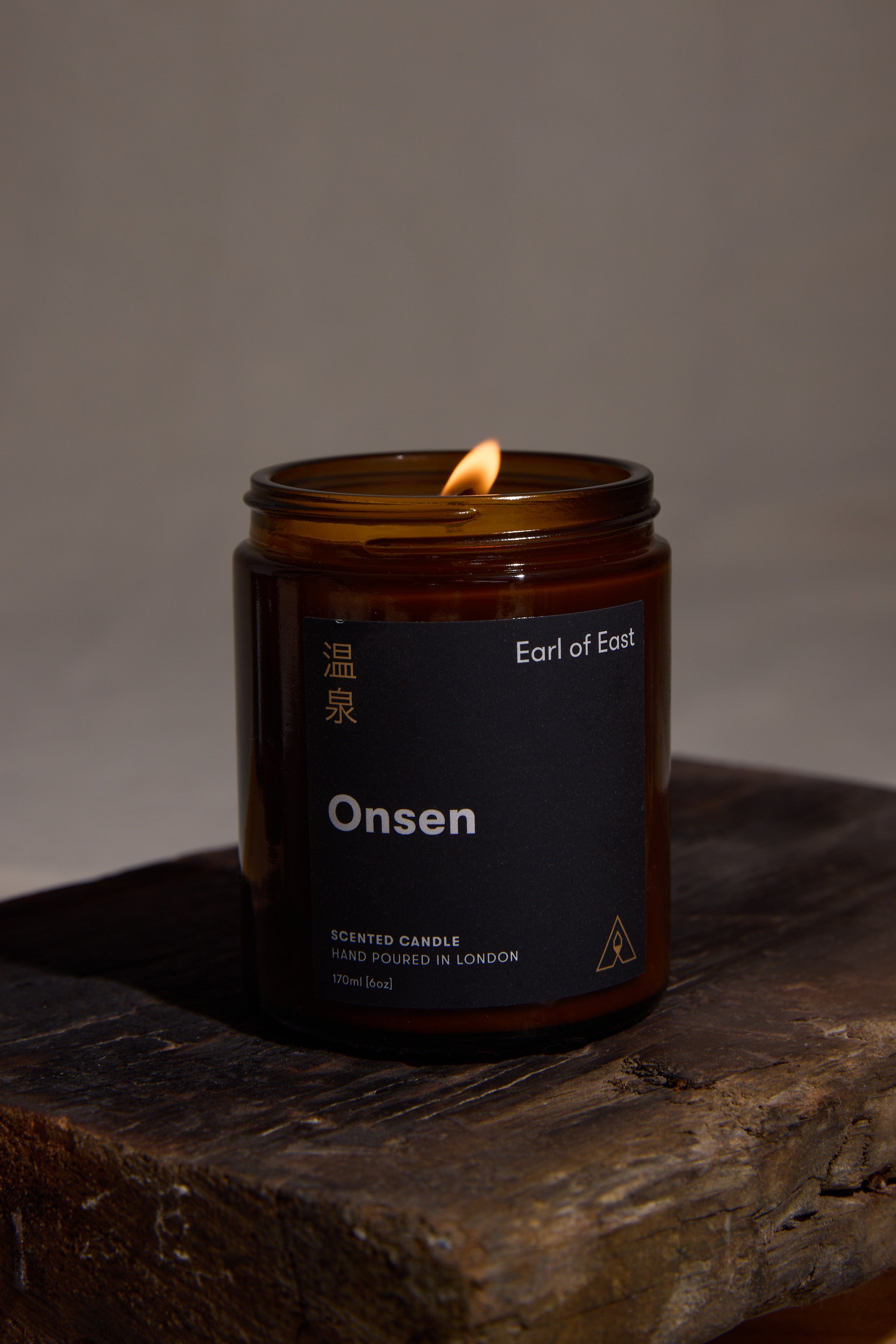 The EoE Onsen Candle 170ml by Earl of East, a peppermint-scented soy wax candle in amber glass with English and Japanese text, rests on rustic wood against a dark backdrop.