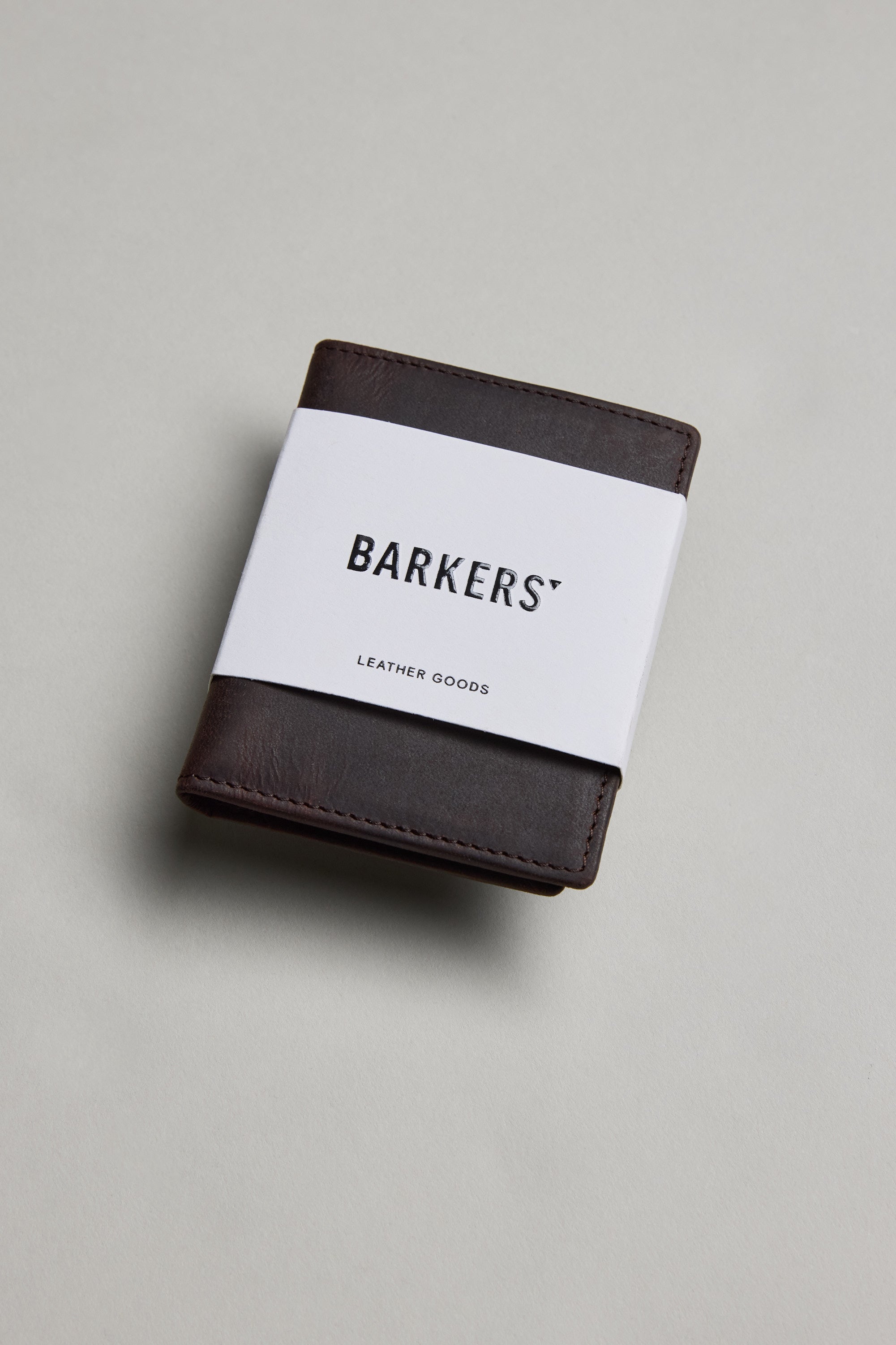 A Marlin Leather Slim Wallet by Barkers, dark brown and crafted from premium cowhide, is wrapped in a white paper band labeled "Barkers" and displayed on a plain light gray surface.