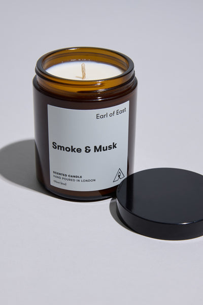 An open EoE Smoke And Musk Candle 170ml by Earl of East rests on a light surface, its black lid beside it. The brown glass jar holds creamy white soy wax and an intact wick, ready to release patchouli notes.