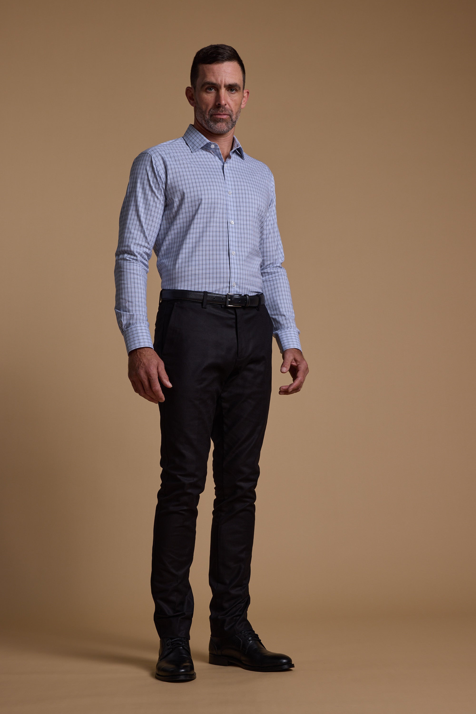 A man with short dark hair, a trimmed beard, and a serious expression wears a Barkers Tabor Check Business Shirt, tailored fit, paired with black dress pants and black shoes against a plain tan background.