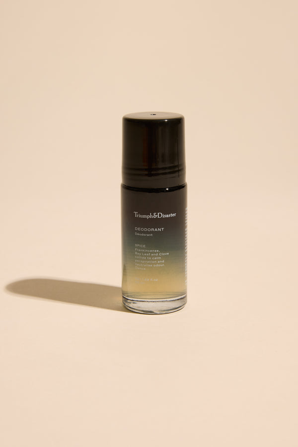 A Triumph & Disaster Deodorant V1 Spice roll-on, featuring a black cap and gradient black-to-clear glass, stands upright on a plain beige background. The label highlights its odour-neutralising organic ingredients.