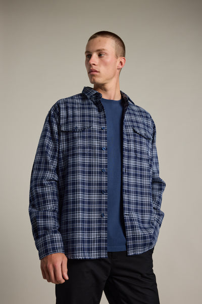 A young man with short hair models the Barkers Jaxon Check Overshirt over a navy t-shirt and black pants, standing with one hand in his pocket and gazing off to the side against a plain background.