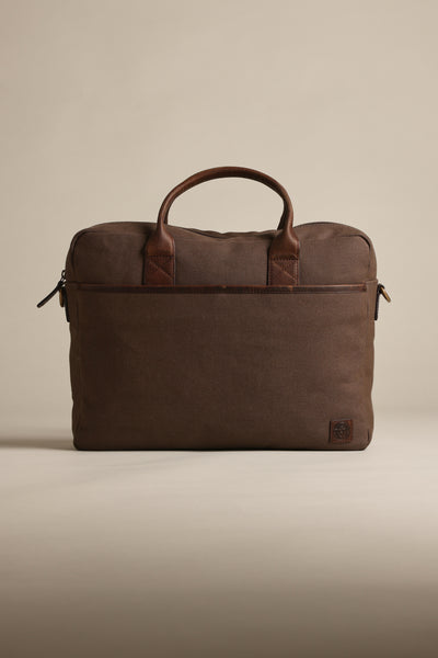 The Barkers Redford Canvas Laptop Bag, featuring brown canvas with premium leather handles and trim, stands upright against a beige background. Its minimalist design includes a zipper closure and a small logo patch in the bottom corner.