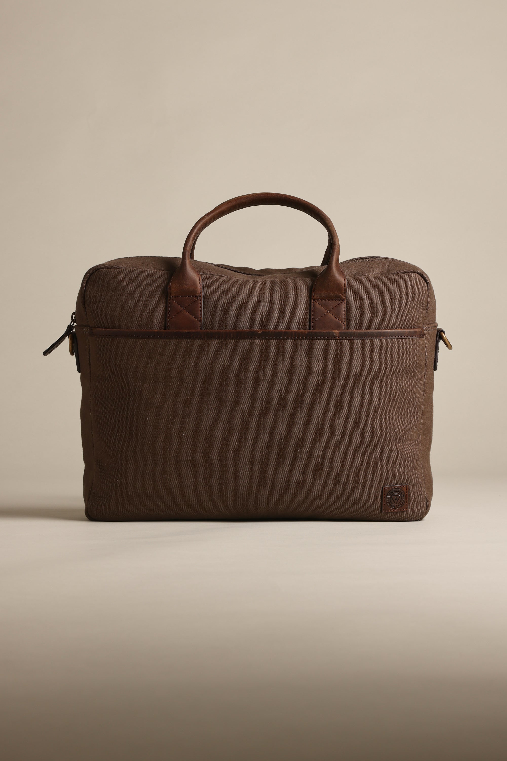 The Barkers Redford Canvas Laptop Bag, featuring brown canvas with premium leather handles and trim, stands upright against a beige background. Its minimalist design includes a zipper closure and a small logo patch in the bottom corner.