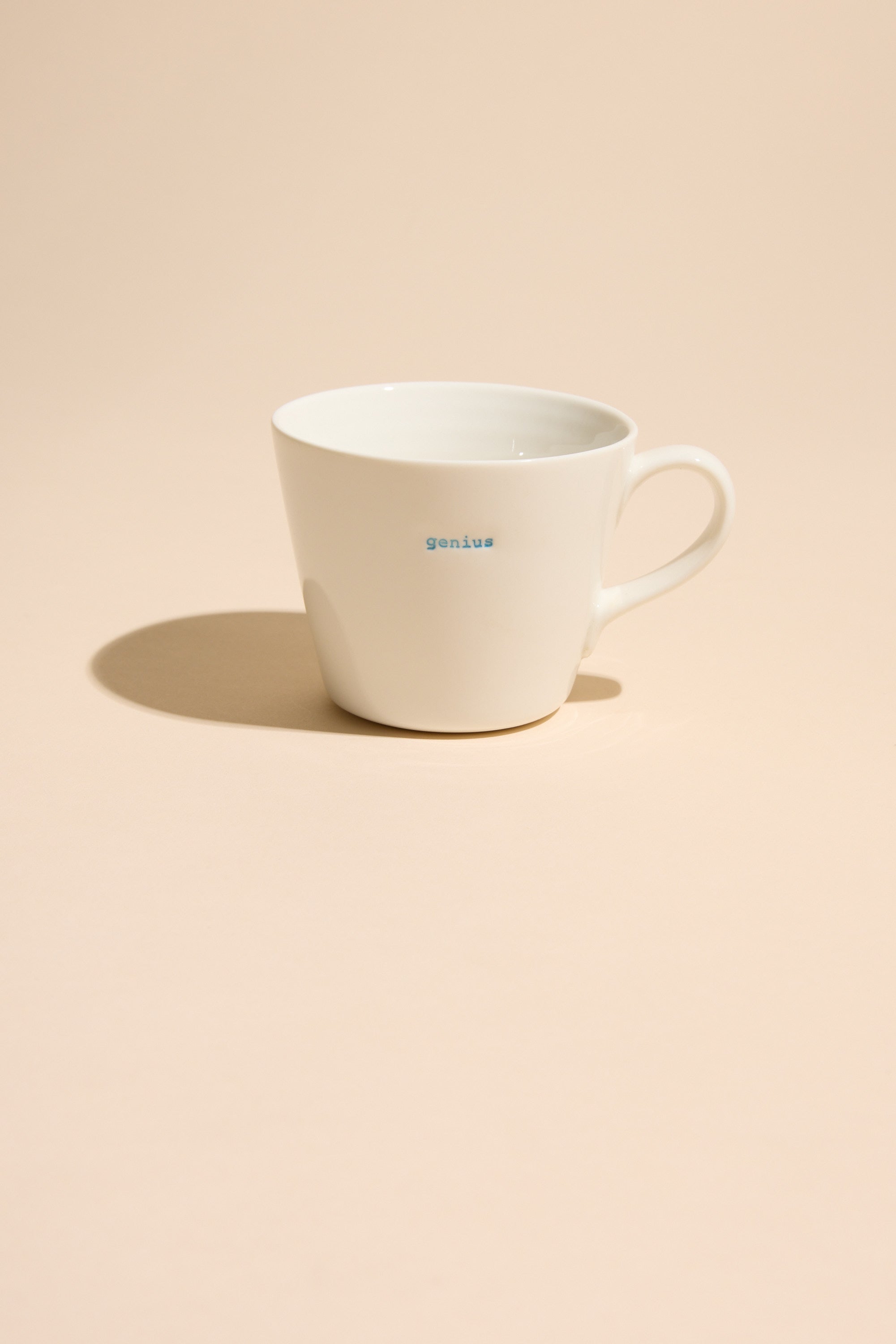 A white ceramic Bucket Mug - Genius by Other, with "genius" in small blue letters, rests on a light beige surface and casts a soft shadow.
