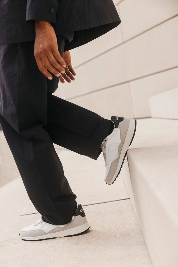 A person in black pants and a black jacket stands on light stone steps, one foot higher. They wear Clae's Joshua sustainable sneakers—white and gray with black soles—crafted from recycled materials by Clae.