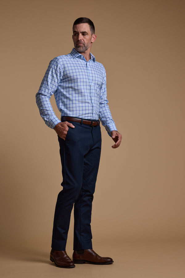 A man stands against a plain beige background wearing the Barkers Bayard Check Business Shirt, paired with dark blue pants, a brown belt, and brown dress shoes. He looks to the side with one hand in his pocket.