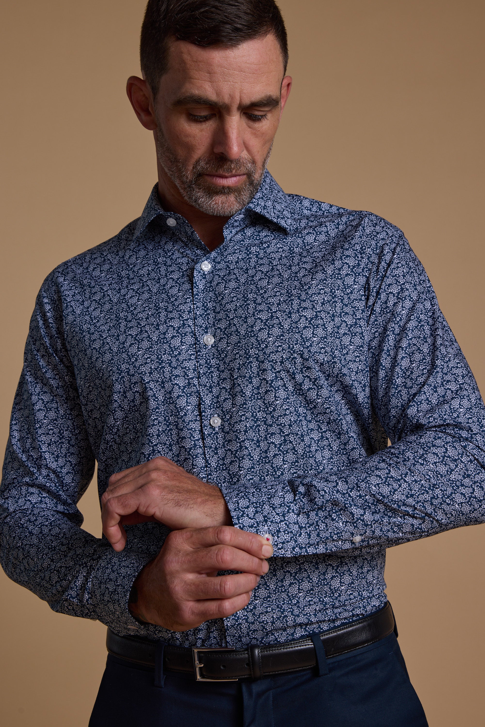 A man in a Barkers Watkins Print Business Shirt, tailored fit, and dark trousers fastens his cuff while looking down. The background is plain beige.
