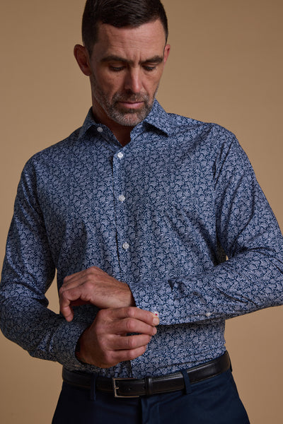 A man in a Barkers Watkins Print Business Shirt, tailored fit, and dark trousers fastens his cuff while looking down. The background is plain beige.