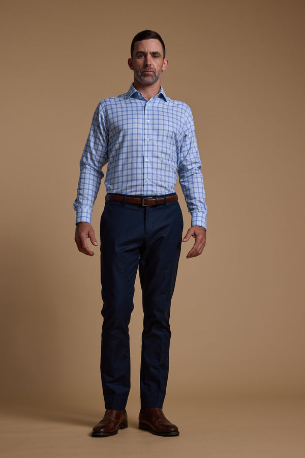 A man faces forward against a plain beige background, wearing the Barkers Bayard Check Business Shirt with Eco Easy Care fabric, dark navy trousers, a brown belt, and brown dress shoes—an ideal business essential. His arms rest relaxed at his sides.