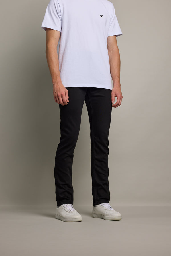 A person, cropped at the shoulders, stands against a plain background wearing Barkers Rambler Denim Slim Jean in black, a white T-shirt, and white sneakers.
