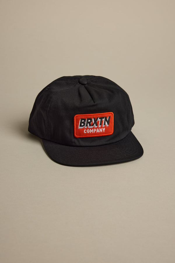 The Brixton Payment MP Snapback is a black adjustable baseball cap featuring a red and white rectangular patch with "BRXTN COMPANY" on the front, shown against a neutral beige background.