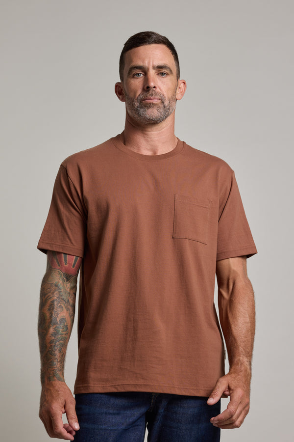 A man with short dark hair, a beard, and a tattooed left arm wears the Barkers Wrenches Print Tee and jeans, standing against a plain light gray background.