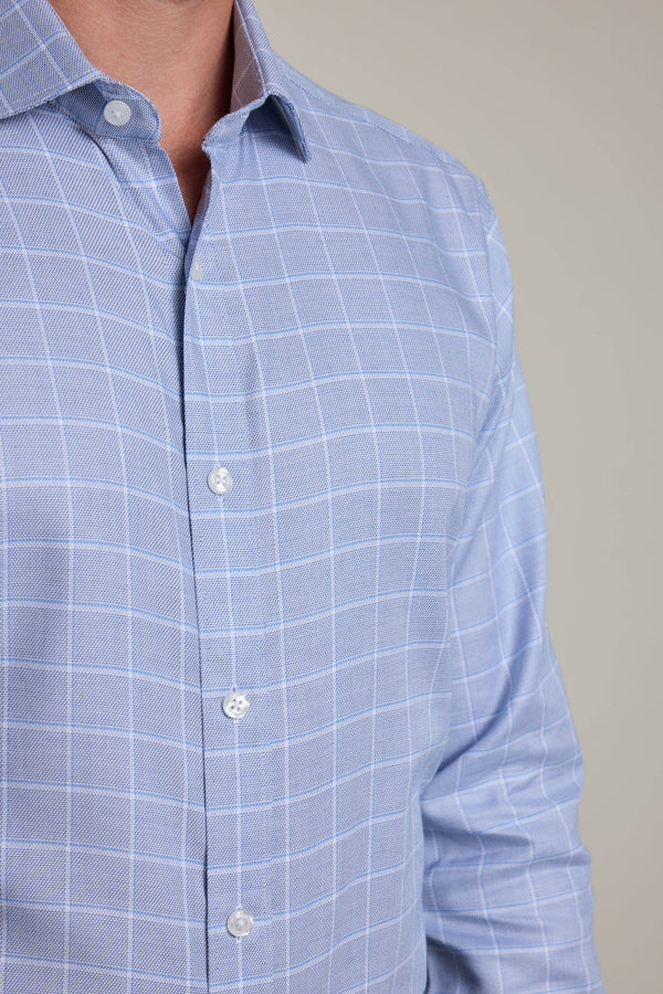 A person wears the Barkers Tyson Check Business Shirt in light blue with a white windowpane check, made from crease-resistant cotton and featuring Eco Easy Care. The shirt is buttoned up against a plain background.