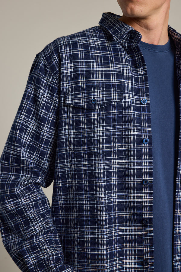 A person wears the Barkers Jaxon Check Overshirt, featuring a vintage-inspired blue plaid pattern, over a plain blue T-shirt. Shown from shoulders to mid-torso, they face slightly to the side.