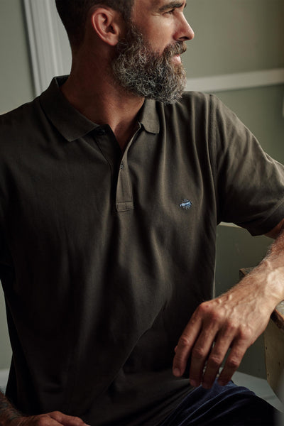 A bearded man wearing a dark Barkers Regent Pique Polo sits indoors near a table, looking to his left with his right arm resting on the table under soft lighting.