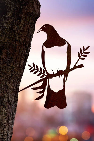 The Metal Birds Kereru / Woodpigeon by Other is a Corten steel bird silhouette holding an olive branch, mounted on a tree trunk with a blurred cityscape and colorful sunset backdrop, inspired by native New Zealand art.