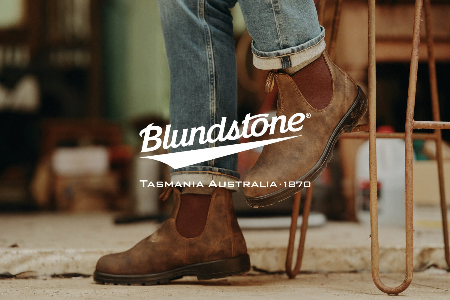 A person wearing brown Blundstone Chelsea boots and blue jeans steps onto a metal bar. The Blundstone logo and Tasmania Australia 1870 are overlaid on the image.