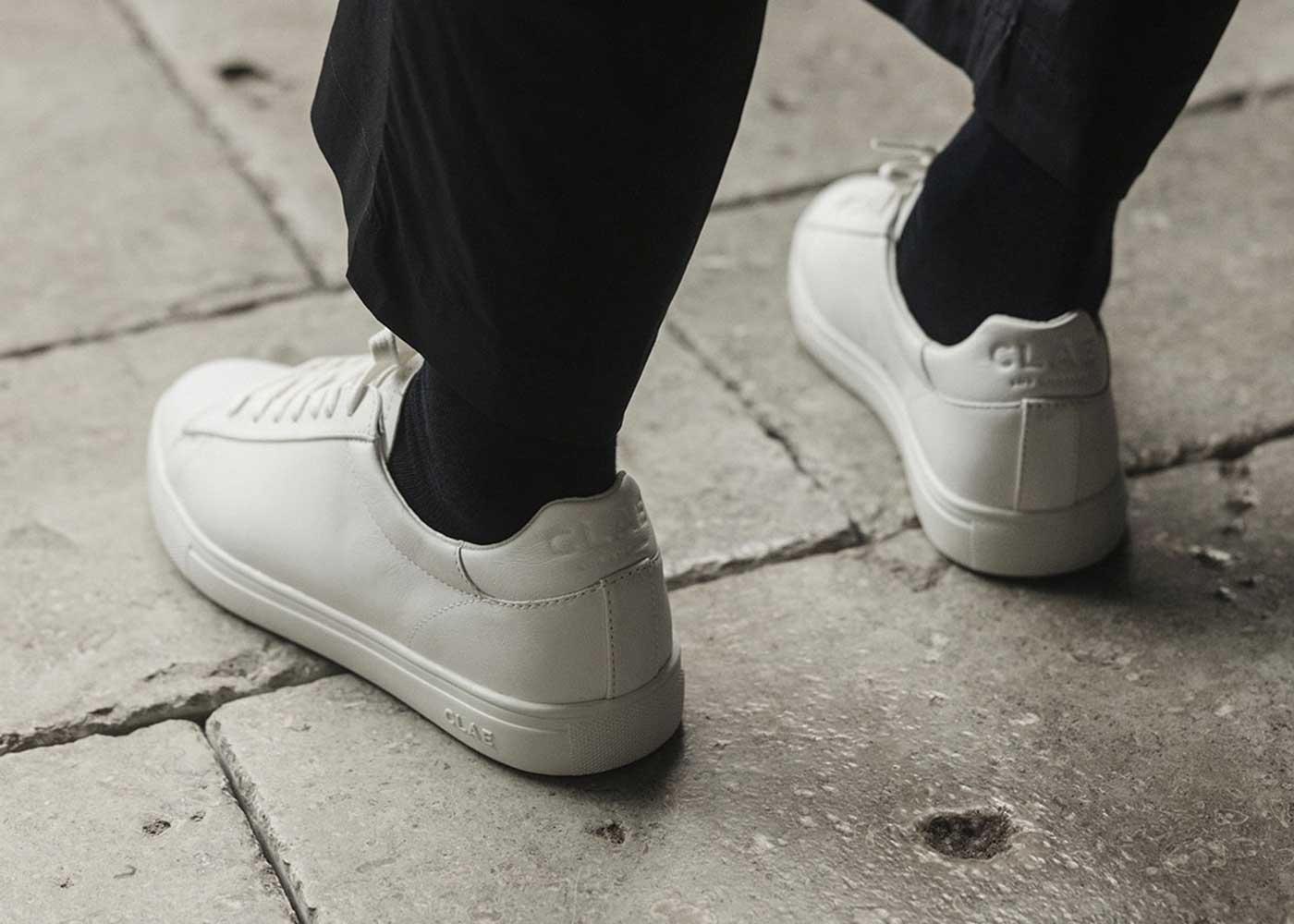 A person wearing black pants and black socks walks on stone pavement, showcasing minimalist white sneakers with a subtle logo on the heel.