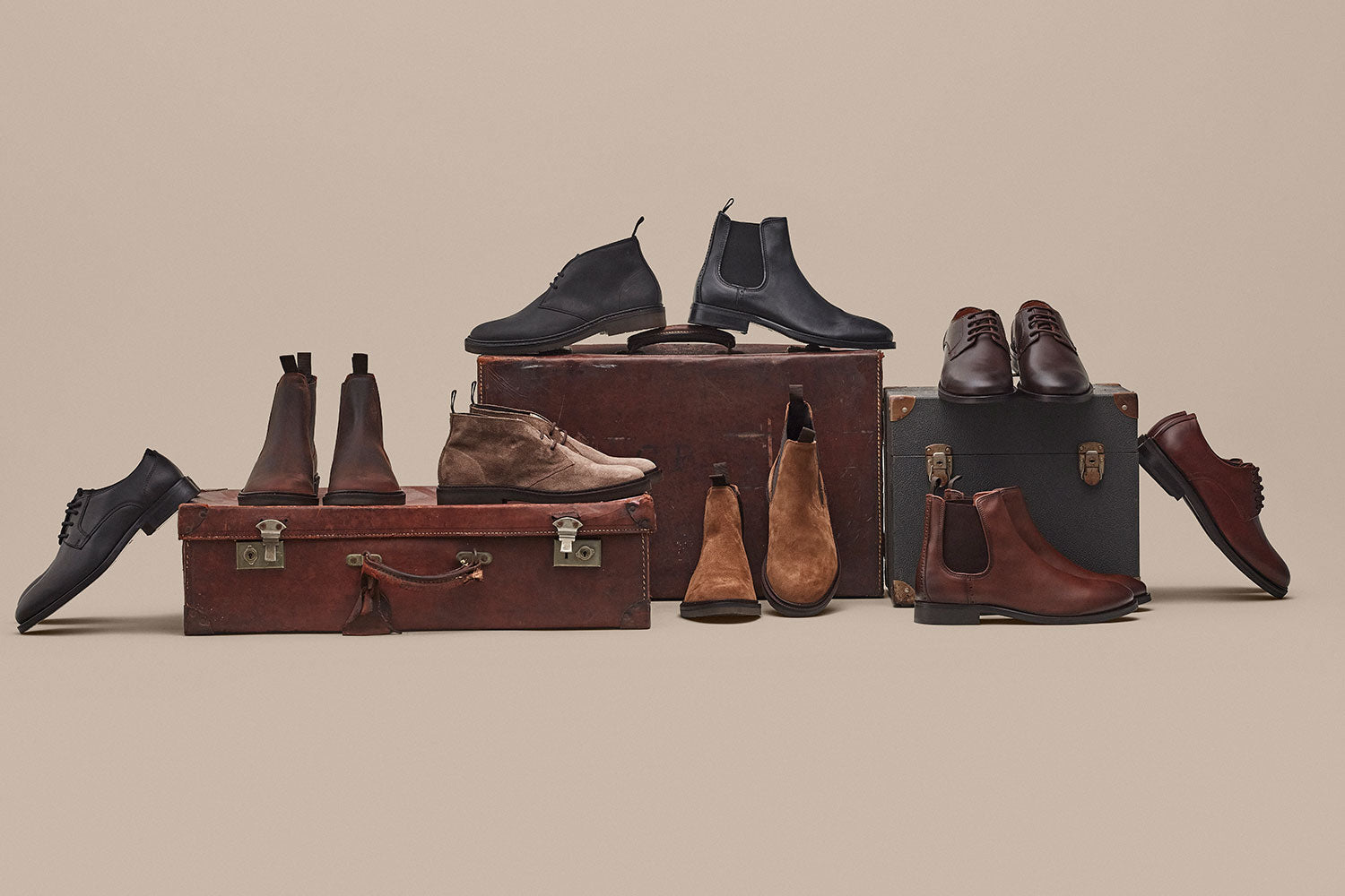 A variety of men’s dress shoes and boots in brown and black are displayed on and around vintage suitcases against a beige background.