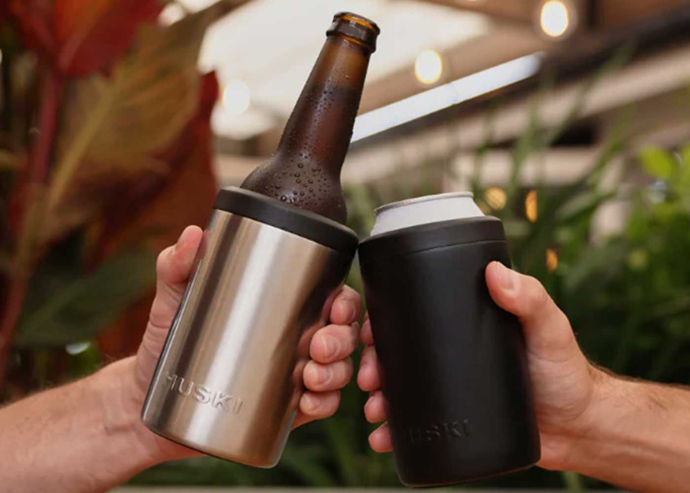 Two hands cheers with drinks in insulated Huski holders—one holding a brown bottle in a silver holder, the other a can in a black holder. Green plants and blurred lights are in the background.