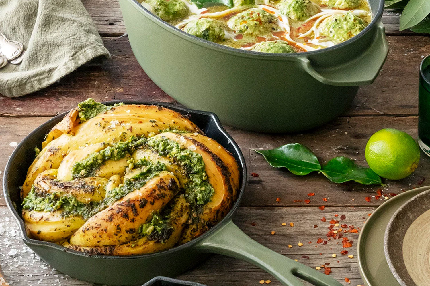 A skillet with golden roasted potatoes topped with green herb sauce sits next to a pot of soup with dumplings, on a rustic wooden table with bay leaves and a lime.