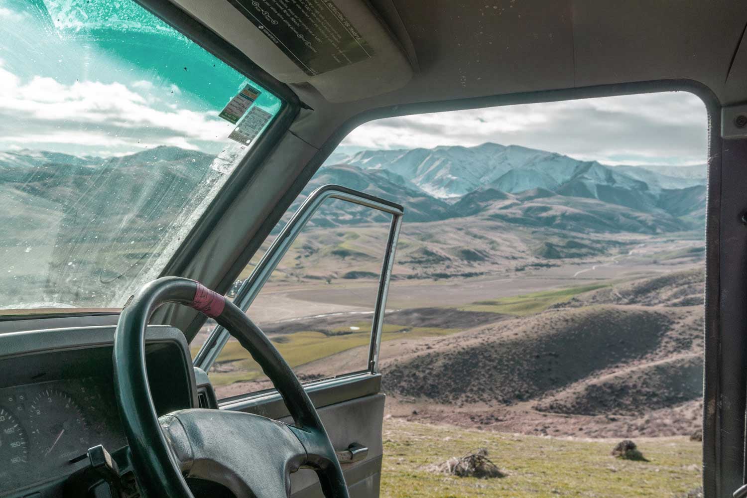 View from inside a vehicle, looking out an open door at a vast, mountainous landscape under a cloudy sky. The steering wheel and dashboard are visible in the foreground.