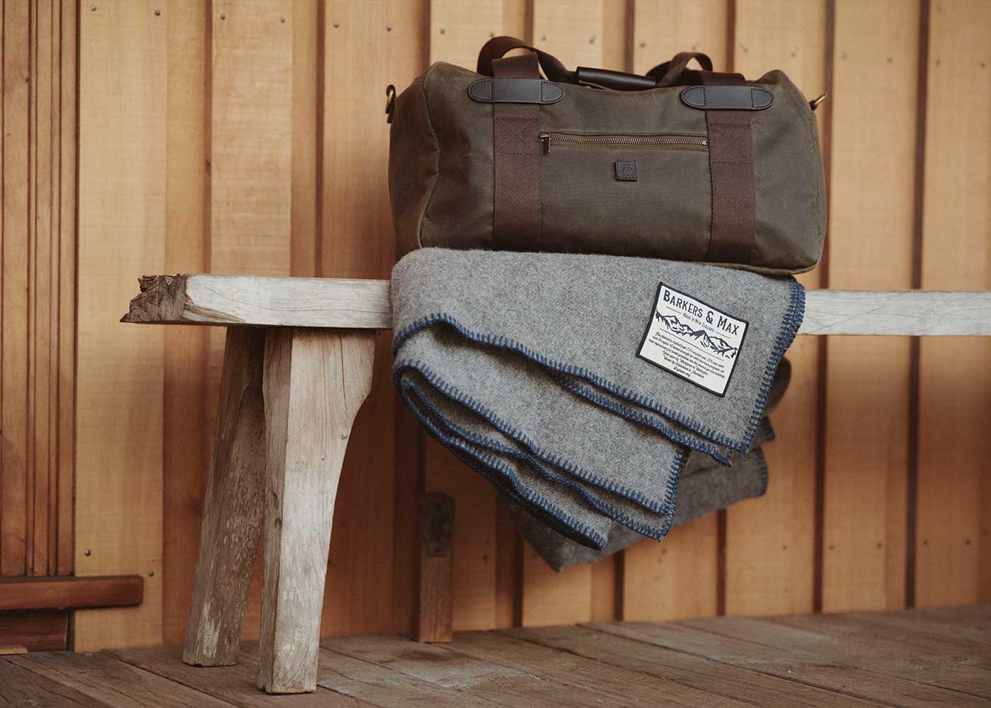 A green duffel bag with brown straps sits on a rustic wooden bench, next to a folded gray wool blanket with blue trim and a visible white label. The background is wooden paneling.