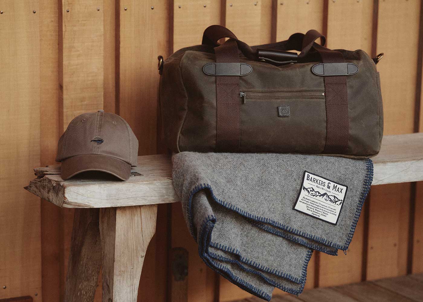 A brown duffel bag, a gray wool blanket with a label, and a brown baseball cap are displayed on a rustic wooden bench against a wooden wall.