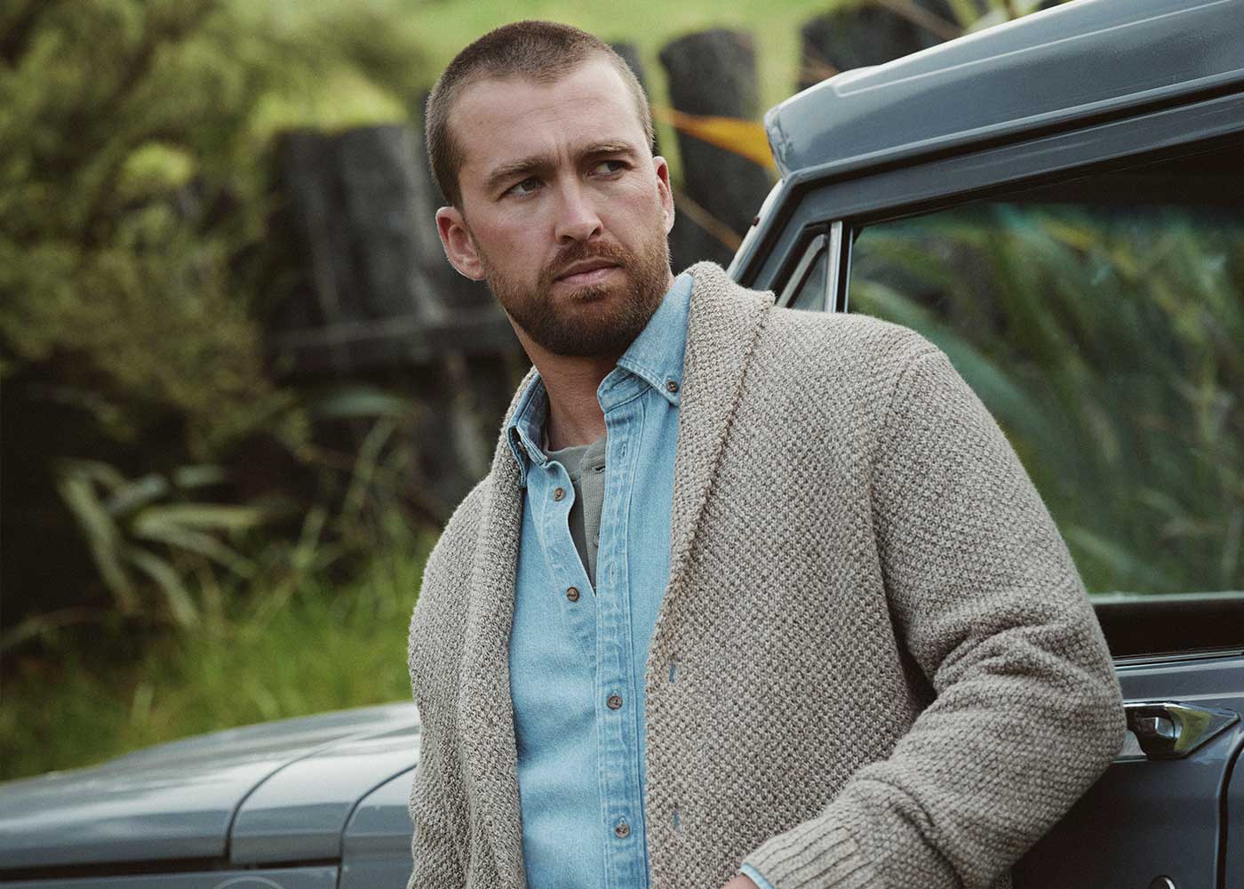 A man with a trimmed beard and short hair, wearing a light blue button-up shirt and a beige textured cardigan, stands beside a vintage gray car outdoors with greenery in the background.