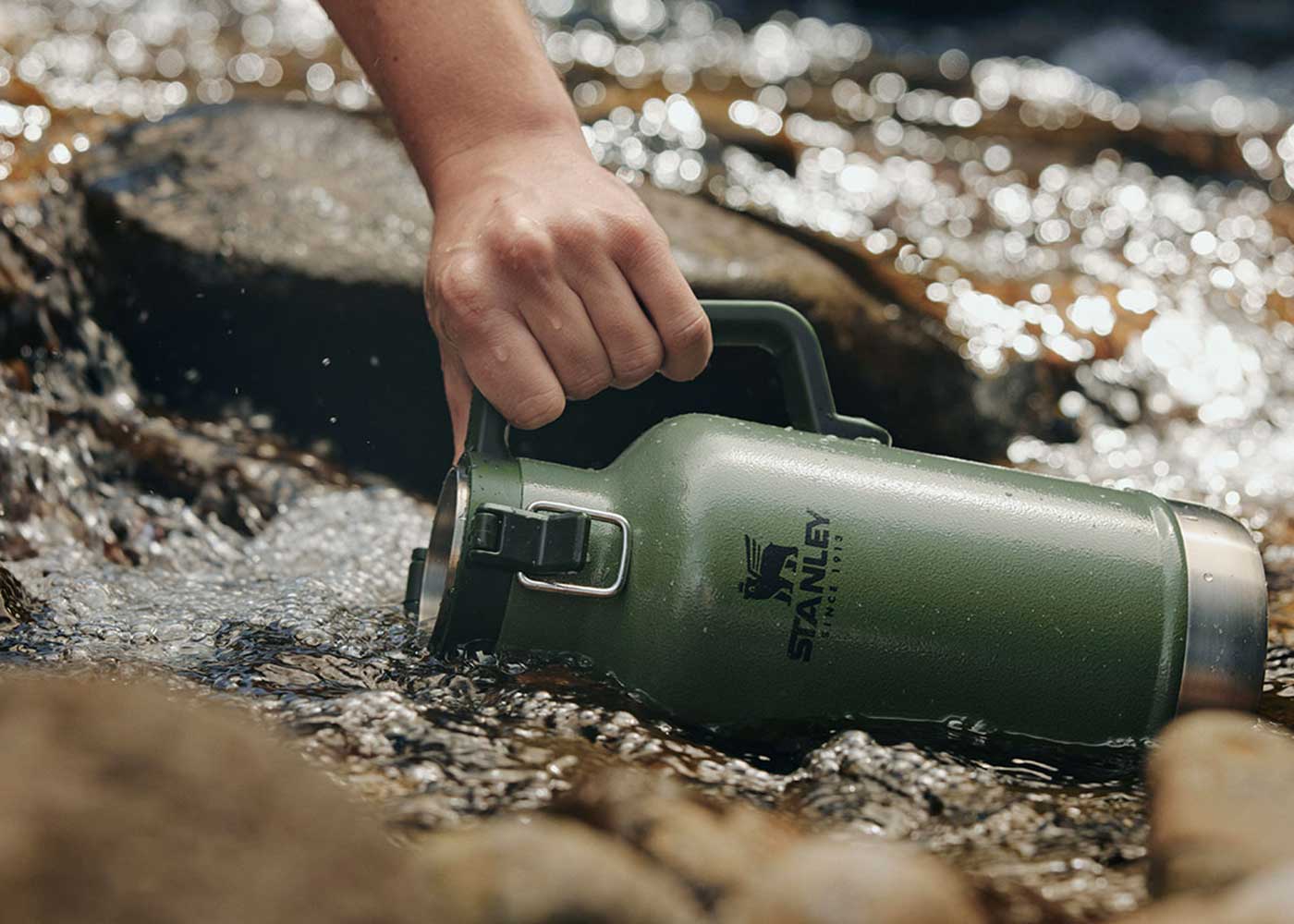 A hand holds a green Stanley thermos, dipping it into a stream among rocks, with water flowing around the bottle.