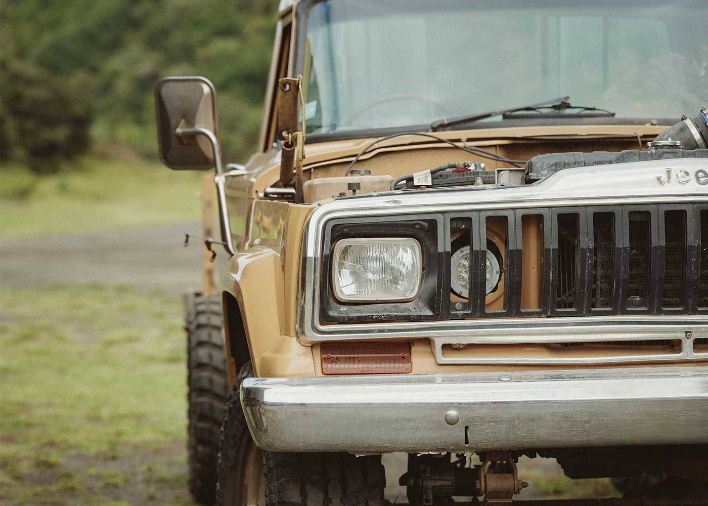 Close-up of the front end of a vintage yellow Jeep truck with its hood open, showing parts of the engine. The vehicle is parked on grass with greenery in the background.