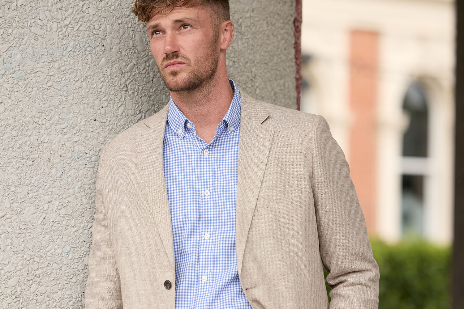 A man in a grey suit and striped tie stands outside a building with turquoise window awnings, looking off into the distance and adjusting his jacket.