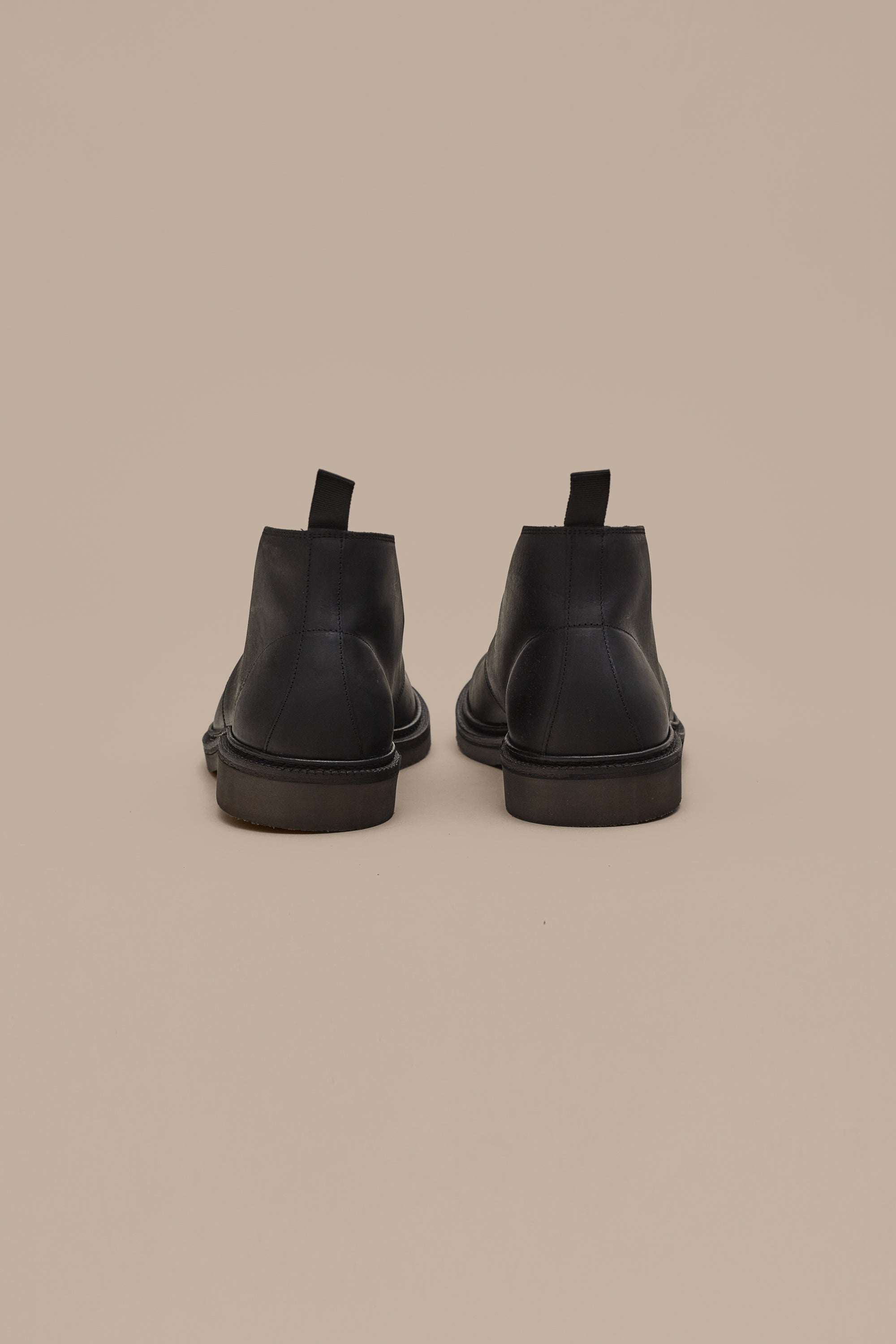 The Barkers Howarth Desert Boot in black leather is displayed from the back, featuring pull tabs on each heel and set against a plain beige background.