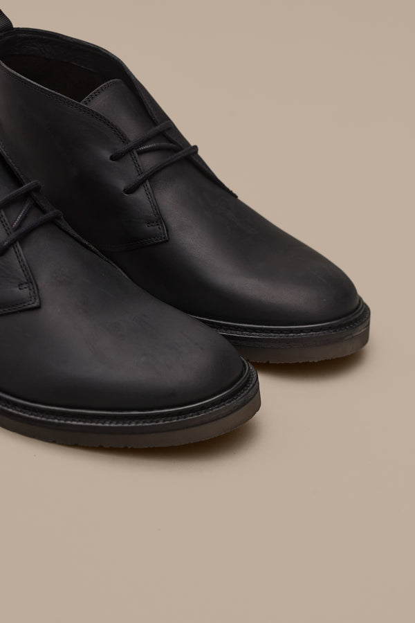 Close-up of the Barkers Howarth Desert Boot in black leather with a smooth, matte finish on a beige surface. Only the front and sides of these stylish lace-up shoes are shown.