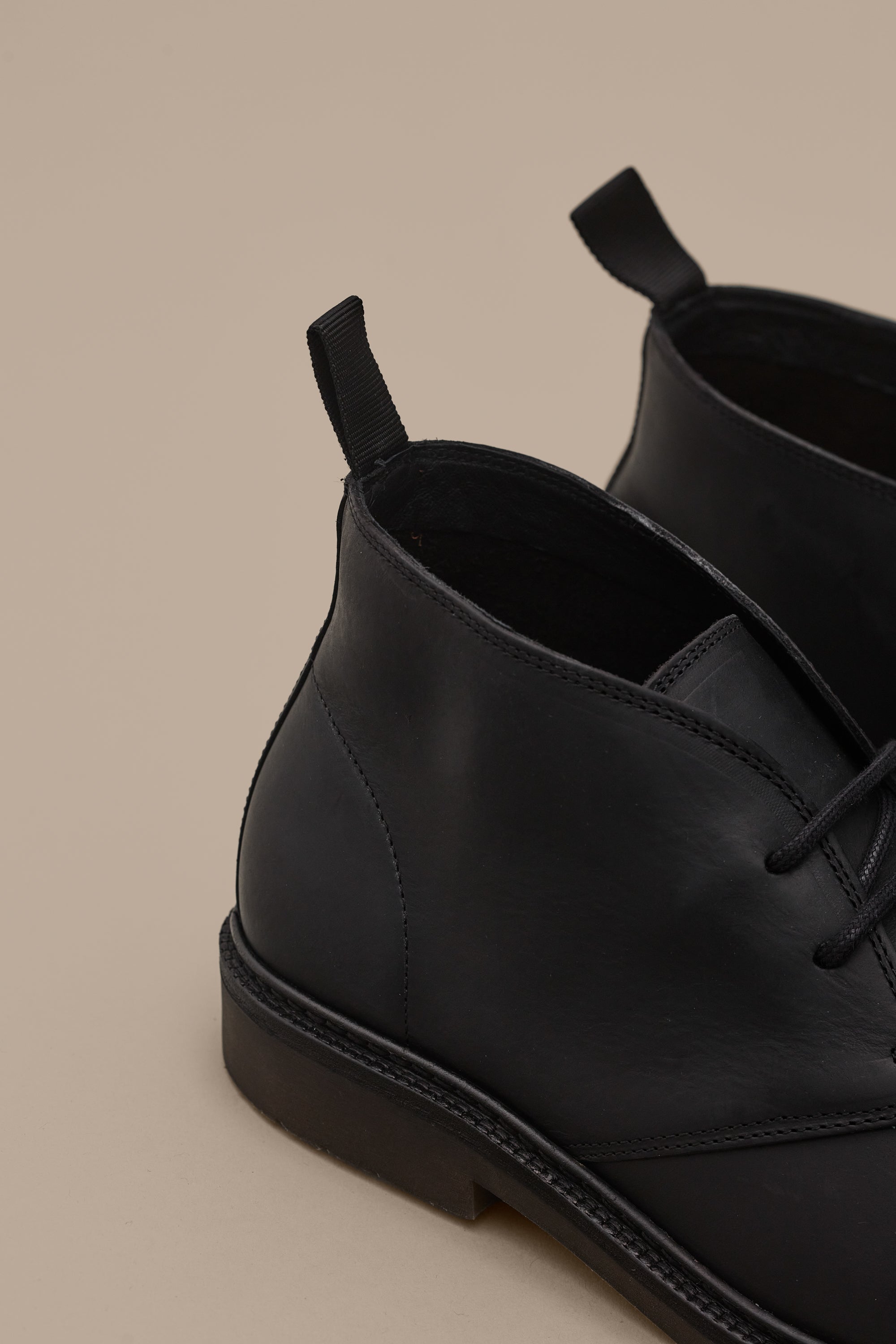 Close-up of Barkers Howarth Desert Boots in black leather with lace-up fronts and pull tabs, shown resting on a beige surface. Only the back and side of these premium Portuguese-made boots are visible.