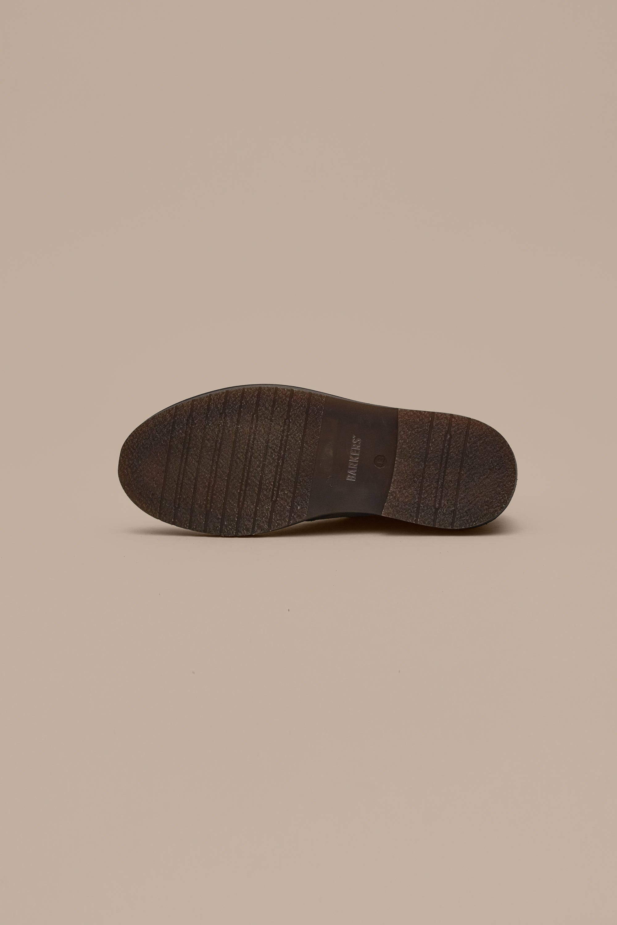 The image displays the bottom view of a Howarth Desert Boot by Barkers, featuring a dark brown sole with horizontal grooves and "Barkers" embossed in the center against a beige background—highlighting quality footwear craftsmanship.