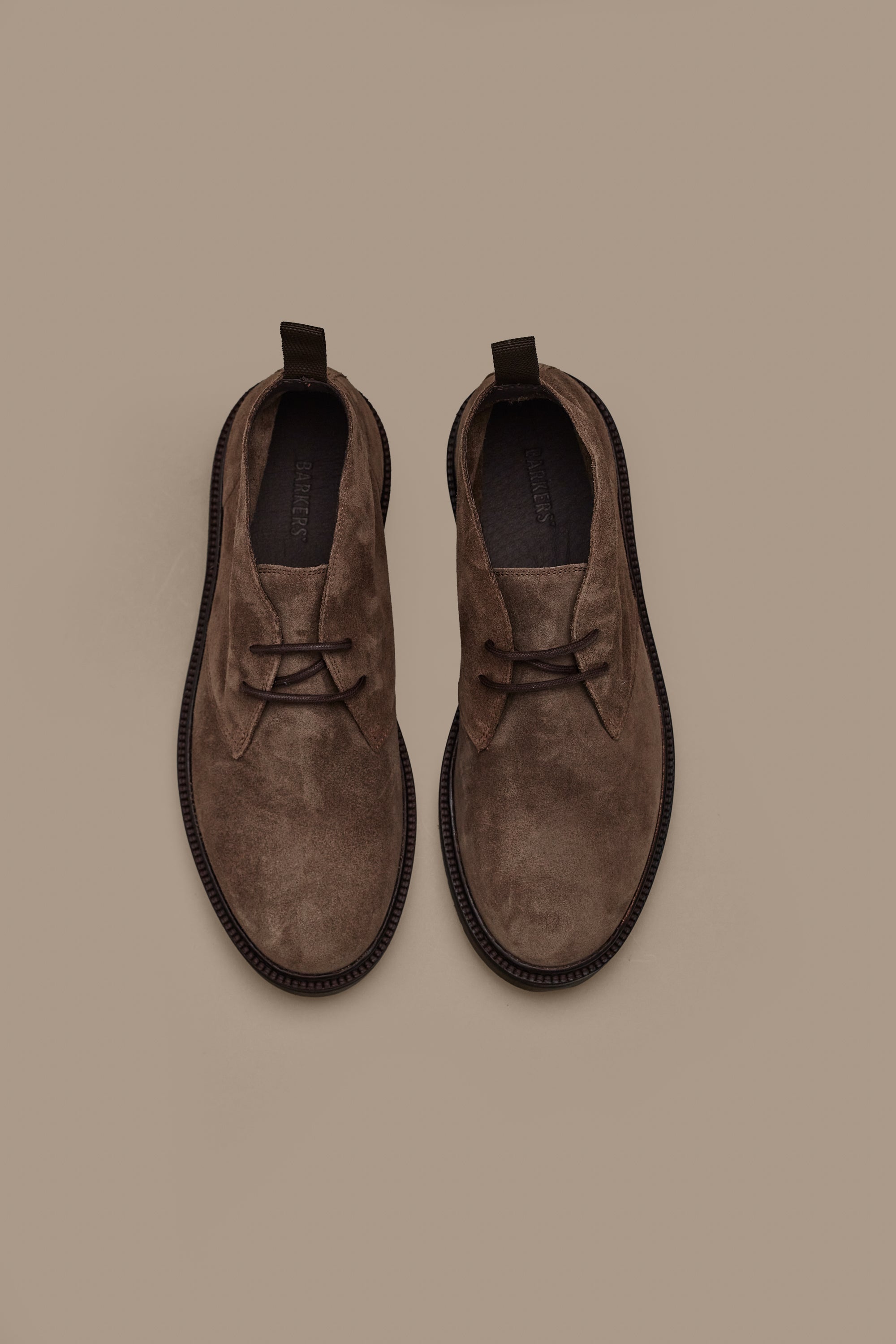 The Barkers Howarth Desert Boot features brown suede, rounded toes, dark soles, and back pull tabs. Displayed side by side from above on a light brown background, these essential boots combine quality and style.