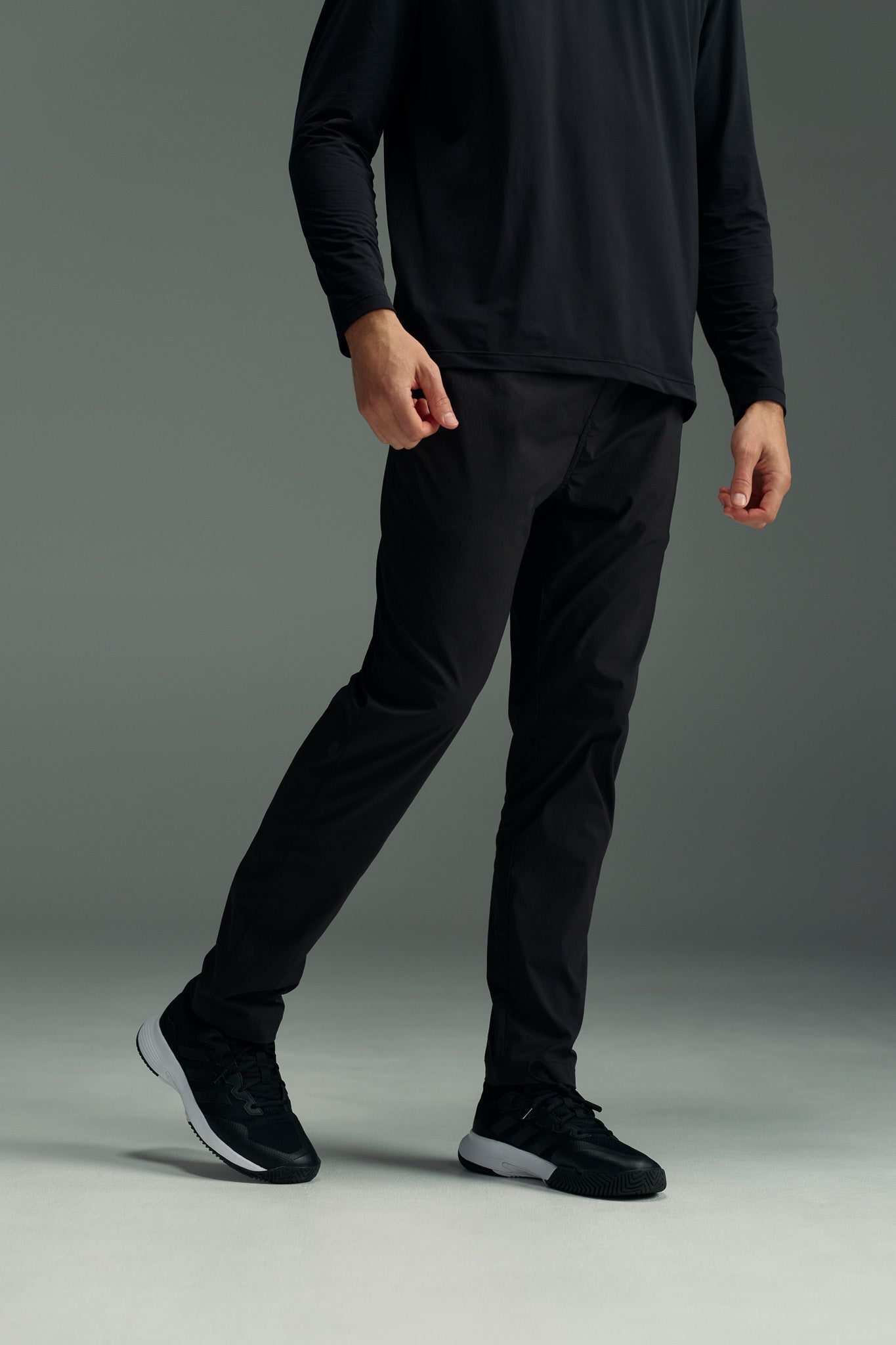A person models the WearEver Transit Pant in black, paired with a long-sleeve black shirt and black sneakers, standing against a plain gray background. Their head is not visible in the image.