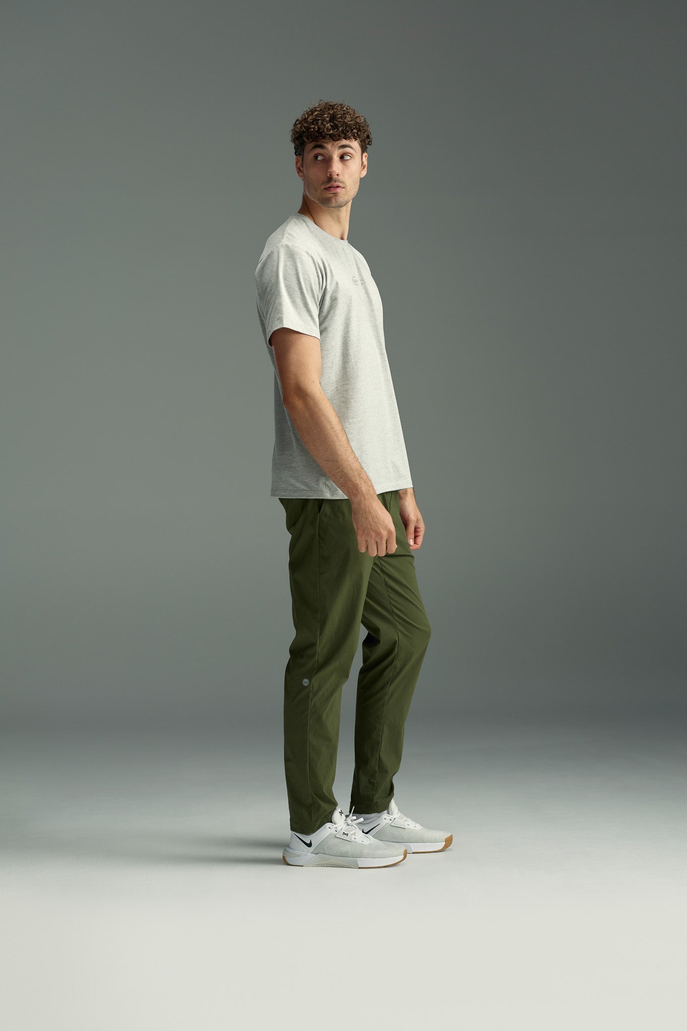 A man with curly hair stands sideways, looking over his shoulder. He wears a light gray T-shirt, white sneakers, and olive green WearEver Transit Pants by WearEver, posed against a plain gray background.