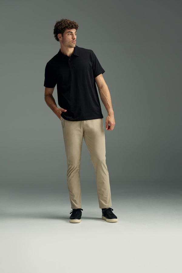 A man with curly hair in a WearEver Outbound Polo, beige pants, and black sneakers stands with one hand in his pocket, gazing to the side against a plain gray background.