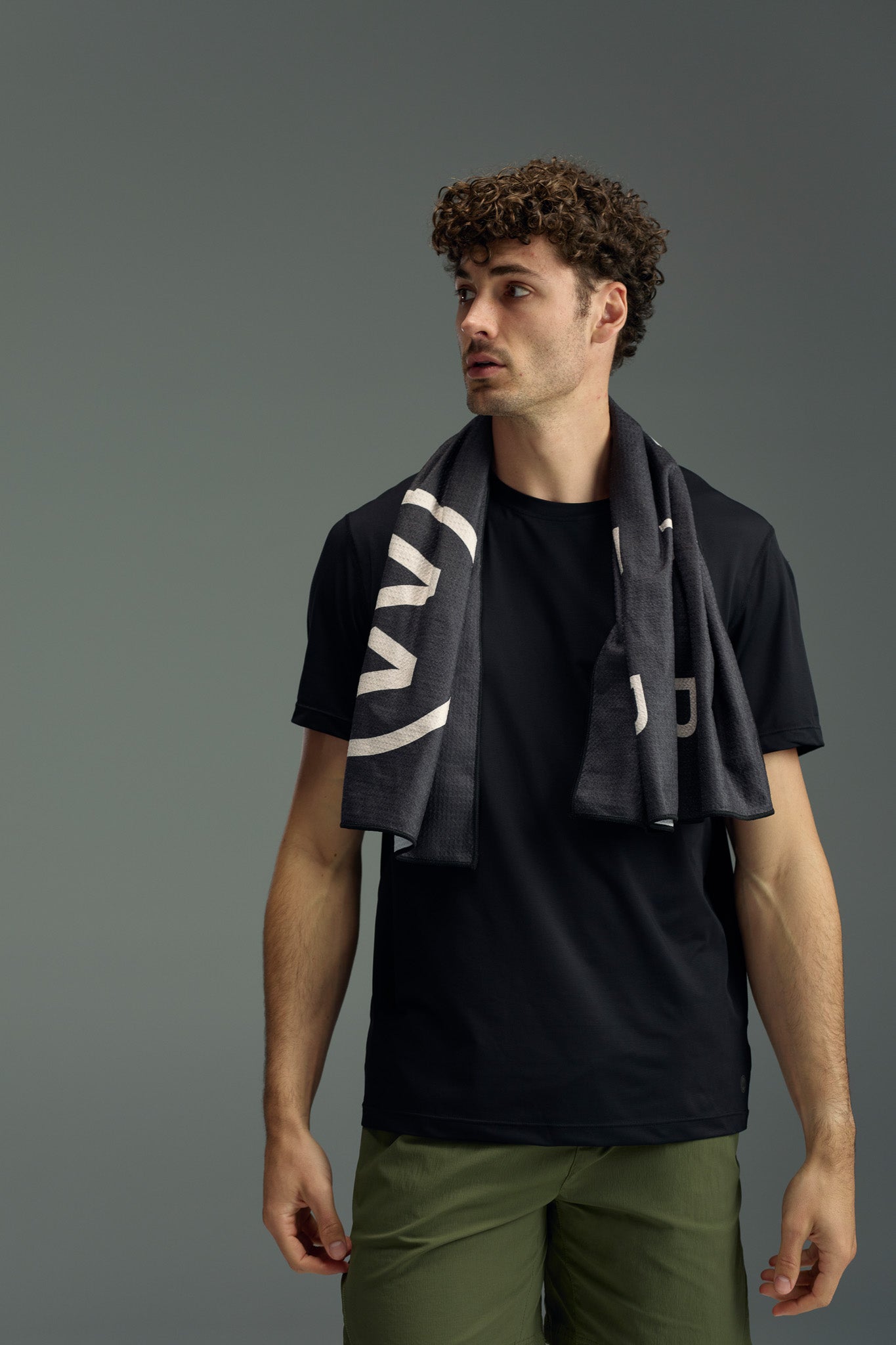 A man with curly hair, dressed in a black t-shirt and olive green pants, stands against a gray background with the WearEver Utility Towel from WearEver draped around his neck, looking to the side.