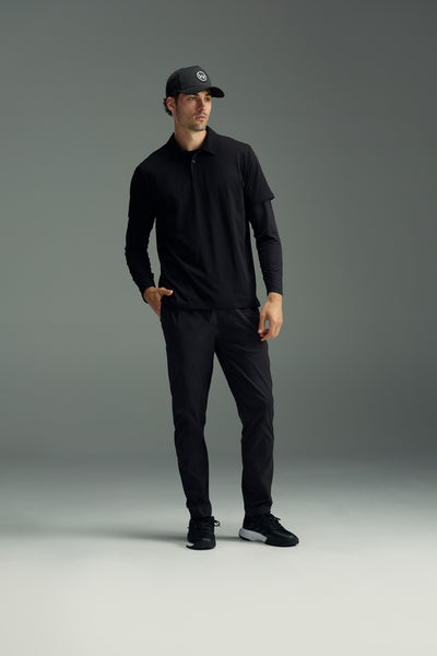 A man in a black baseball cap, black polo shirt, black long sleeves, WearEver Transit Pant by WearEver, and black sneakers stands with one hand in his pocket, looking off to the side against a plain gray background.