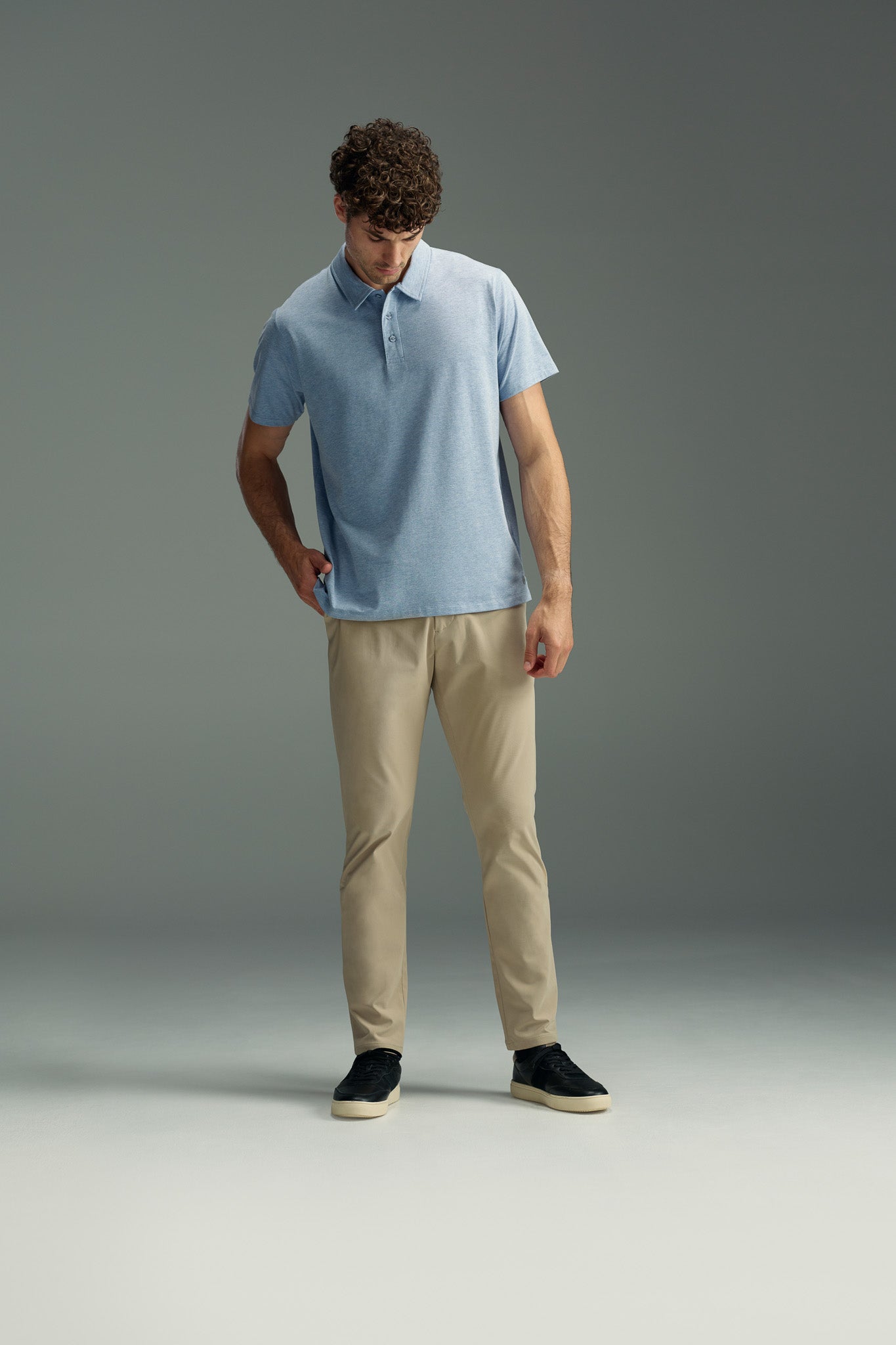 A man in a light blue WearEver Outbound Polo by WearEver, crafted from breathable cotton-blend fabric, pairs it with beige pants and black sneakers as he stands against a gray background, looking down with one hand in his pocket.