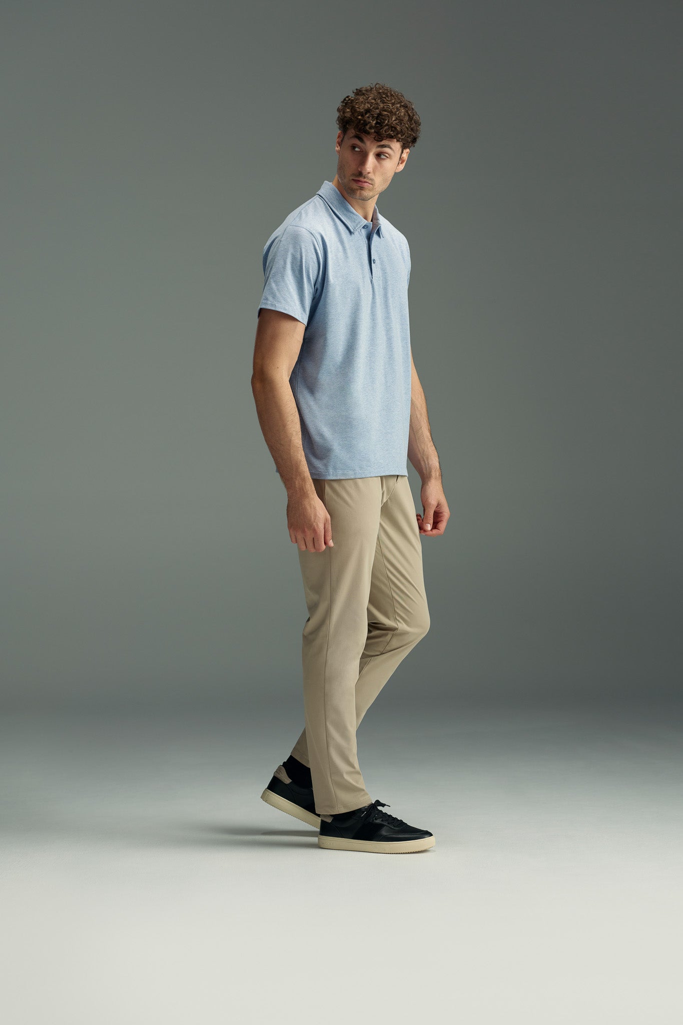 A man with curly hair wears a light blue WearEver Outbound Polo from WearEver, made of breathable cotton-blend fabric, along with beige pants and black sneakers. He stands against a plain gray background, looking off to the side.