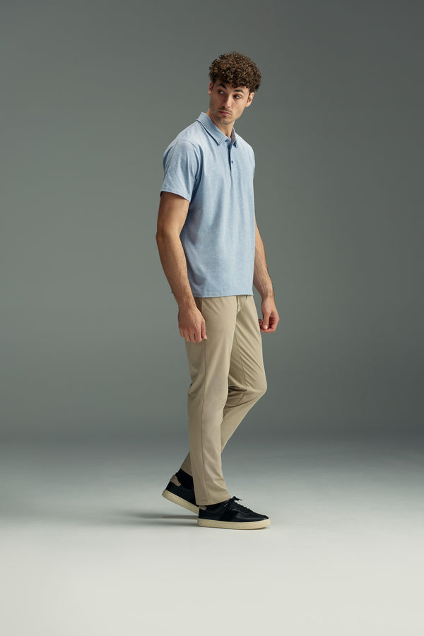 A man with curly hair wears a light blue WearEver Outbound Polo from WearEver, made of breathable cotton-blend fabric, along with beige pants and black sneakers. He stands against a plain gray background, looking off to the side.