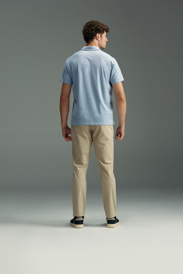 A man with curly hair wears a light blue, breathable cotton-blend WearEver Outbound Polo, beige pants, and black shoes while facing away from the camera against a plain gray background.